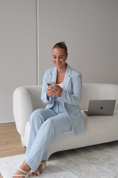 a woman sitting on a white couch holding a cell phone and looking at her laptop