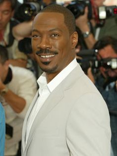 a man in a white suit posing for the camera with cameras around him and people behind him