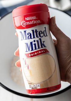 a hand holding a bottle of malted milk next to a plate with food on it