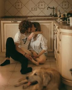 two people sitting on the floor in a kitchen with a dog laying next to them