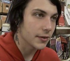 a man with black hair wearing a red hoodie in front of bookshelves