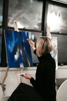 a woman sitting in front of a easel holding a paintbrush and painting on an easel