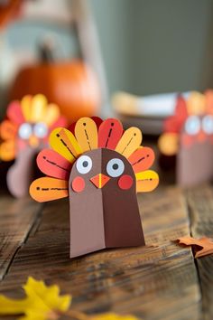 two paper turkeys sitting on top of a wooden table next to orange and yellow leaves