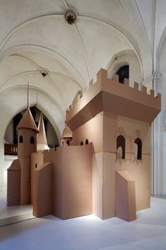 a castle made out of cardboard sitting inside of a white walled building with arched windows