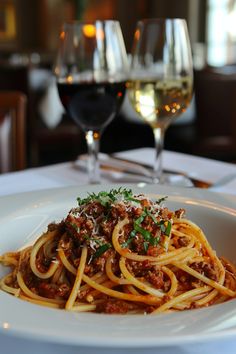 a white plate topped with spaghetti and sauce next to two glasses of wine on a table