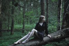 a woman sitting on top of a fallen tree in the woods with her legs crossed