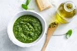 a white bowl filled with green pesto next to a wooden spoon and bottle of olive oil