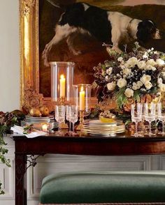 a table with candles, flowers and plates on it in front of a large painting