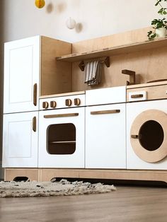 a toy kitchen with white appliances and wooden cabinets on the floor next to a potted plant