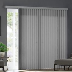 a living room with white vertical blinds in the window, and a grey chair next to it