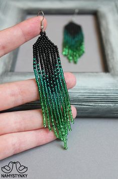 a hand holding a pair of green and black beaded fringe earrings in front of a frame