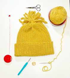 a yellow knitted hat next to a ball of yarn and knitting needles on a white surface