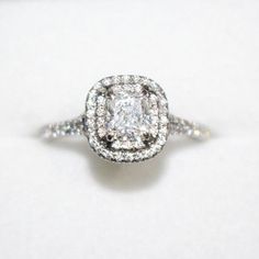 an engagement ring with a square diamond center surrounded by pave diamonds on a white surface