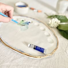 a person is painting on a plate with paintbrushes and some white flowers in the background