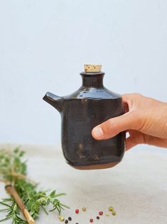 a hand is holding a black teapot on a table with herbs and spices around it