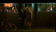 a man and woman standing in front of a store at night with neon lights on the windows