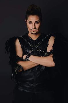 a man with his arms crossed wearing black leather and feathers on his chest, standing in front of a dark background