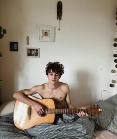 a shirtless man sitting on a bed with an acoustic guitar in his hand and looking at the camera