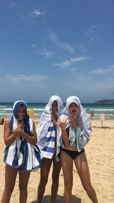 three women in bathing suits standing on the beach with towels around their necks and smiling