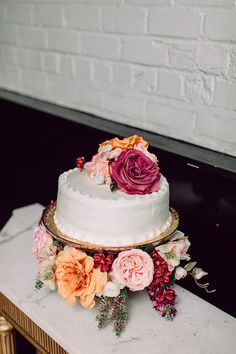 a three layer cake with flowers on top sitting on a table next to a brick wall