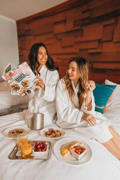 two women in white robes sitting on a bed with breakfast foods and drinks around them