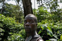 a young man standing in front of some trees and bushes looking up at the camera