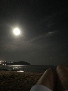 a person sitting on the beach at night watching the moon rise over the water and land