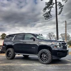 a black truck parked in a parking lot next to a tall pine tree on a cloudy day