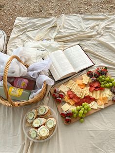 an open book and some snacks on a blanket with a basket full of cheeses