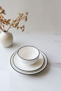 three white plates with black rims sit on a marble surface next to a flower vase