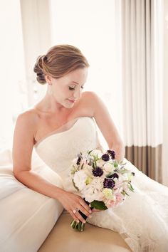 a woman in a wedding dress sitting on a couch holding a bridal bouquet with purple and white flowers