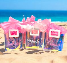 three bags with pink bows are on the sand near the ocean and blue sky in the background