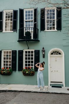 a woman standing in front of a green building with black shutters and windows on the side