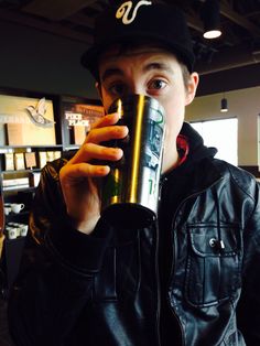 a man in a leather jacket drinking from a beer can while wearing a black hat