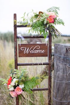 a wooden welcome sign sitting on top of a field next to a fence with flowers and greenery