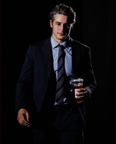 a man wearing a suit and tie holding a drink in his hand while standing against a black background