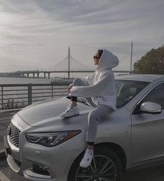 a man sitting on the hood of a silver sports car in front of a bridge