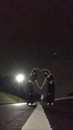 two young women standing on the side of a road at night with their arms in the shape of a heart