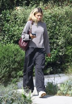 a woman in grey sweater and black pants walking down the street with her cell phone