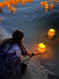 a woman kneeling down next to water with floating lanterns in the air above her head