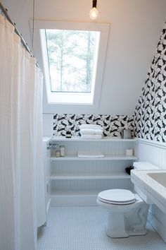 a white toilet sitting under a bathroom window next to a sink and bathtub covered in black and white wallpaper