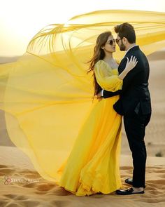 a man and woman standing in the sand with their arms around each other, wearing yellow dresses