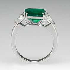 This captivating ring is centered with one (1) emerald cut natural emerald set into a four-split prong setting. The center stone is flanked to each side by one (1) triangular modified brilliant cut diamond set into a three-prong setting. The ring measures 10.9mm at the top, rises 7.4mm above the finger, tapering to 2.6mm wide and 1.5mm thick at the base of the shank. This ring is currently a size 8.25. Formal Radiant Cut Emerald Ring With Bezel Setting, Oval Three Stone Emerald Ring For Formal Occasions, Formal Emerald Ring With Radiant Cut For May Birthstone, Formal Emerald Three-stone Ring, Formal Emerald Ring With Center Stone Octagon Shape, Formal Octagon Emerald Ring With Center Stone, Formal Emerald Ring With Radiant Cut, Classic Trillion Cut Emerald Ring For Formal Occasions, Fine Jewelry Emerald Ring In Radiant Cut Platinum