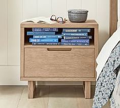 a nightstand with books on top of it next to a bowl and eyeglasses