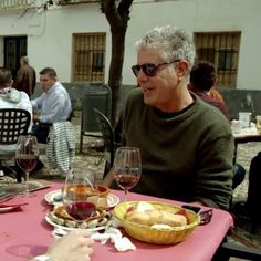 two men sitting at a table with food and wine in front of them, talking to each other