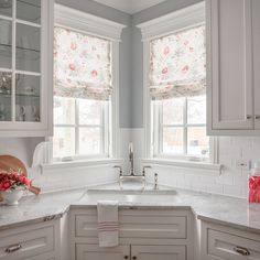 a kitchen with white cabinets and marble counter tops in front of two windows that have floral roman shades on them