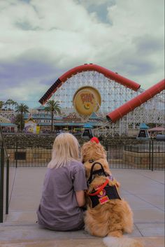 a woman sitting on the ground next to a dog in front of a roller coaster