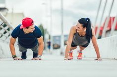 a man and woman doing push ups on the sidewalk together, one is wearing a red hat