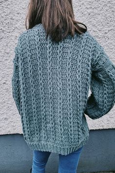 a woman standing in front of a wall wearing a green knitted sweater and jeans