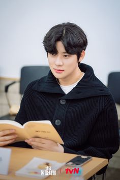 a young man sitting at a table reading a book while wearing a black coat and white shirt